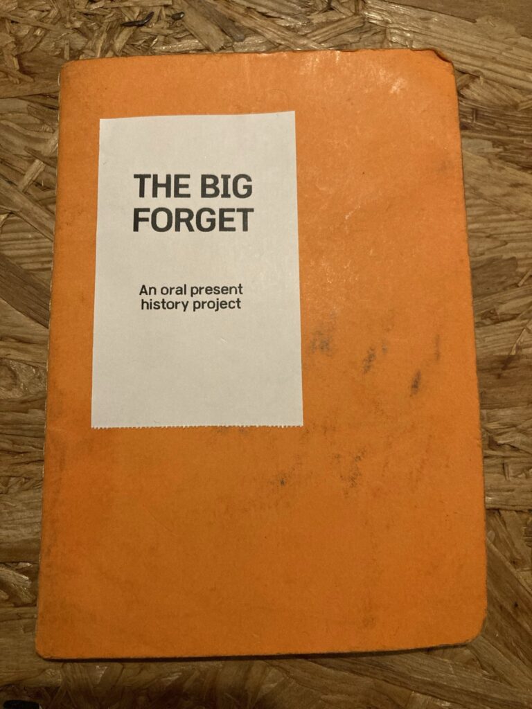 A small orange notebook against a wooden floor. A white sticker. Black text reads The Big Forget, an oral present history project 